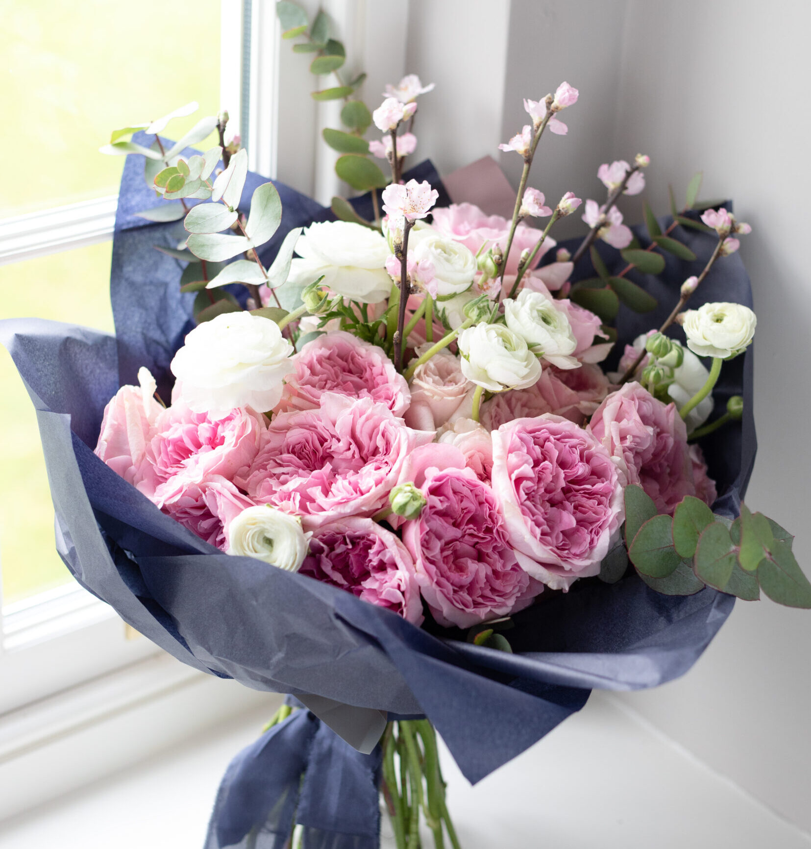 Luxury hand-tied gift bouquet featuring pink garden roses, seasonal flowers and foliage wrapped in dark paper.